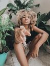 Unemotional female with frizzy hair in summer clothing confidently looking at camera while resting in bright room decorated with pot plants