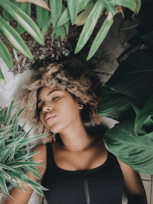 Overhead of gentle black woman with blond Afro hair resting on floor with eyes closed surrounded by pot flowers