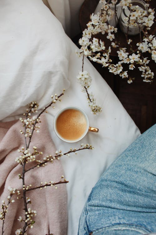 Top view of anonymous female sitting on bed with mug of fresh cappuccino and flowers while enjoying cozy morning
