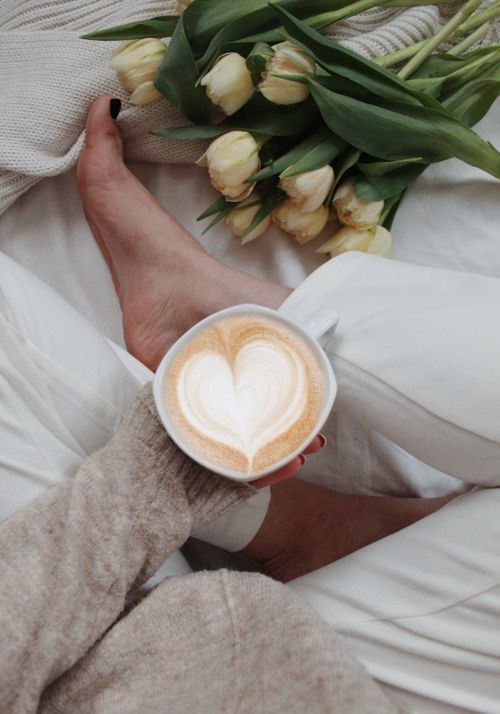 Free From above of unrecognizable female with cup of cappuccino with heart shaped foam sitting on bed with flowers in morning Stock Photo