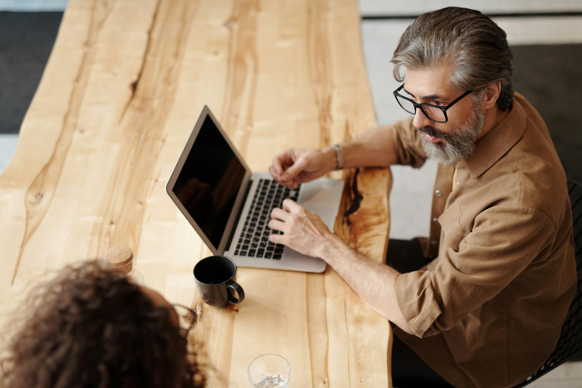 Adult man with beard and eyeglasses typing on laptop at wooden desk indoors.