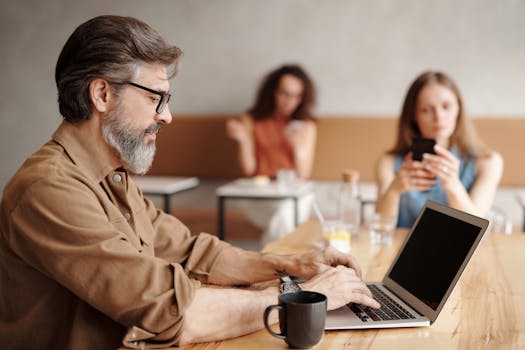 Mature man with laptop working in a coffee shop, surrounded by a busy environment.