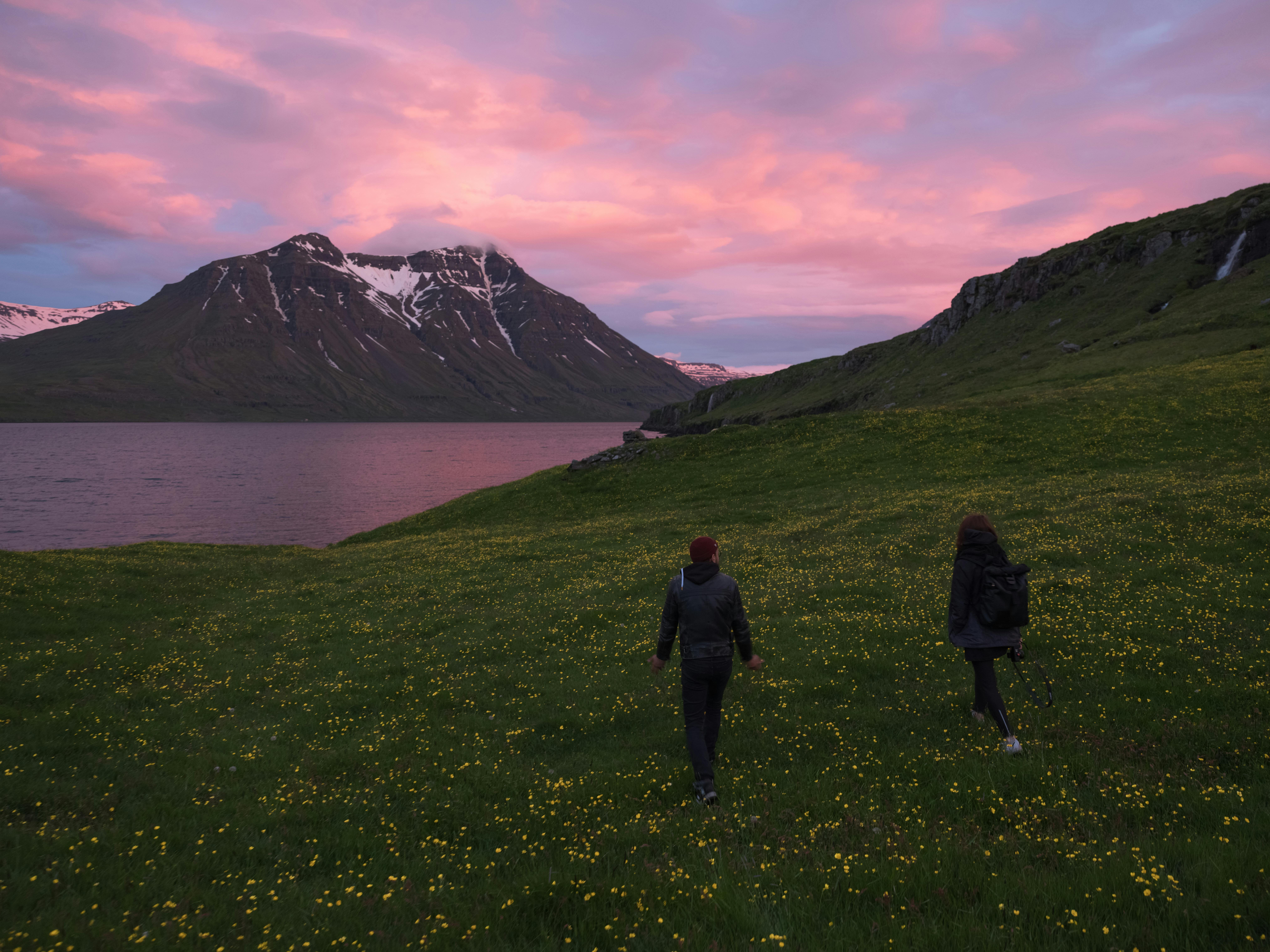 Back view full length unrecognizable tourists walking on grassy hill towards river and majestic mountain range under purple sky