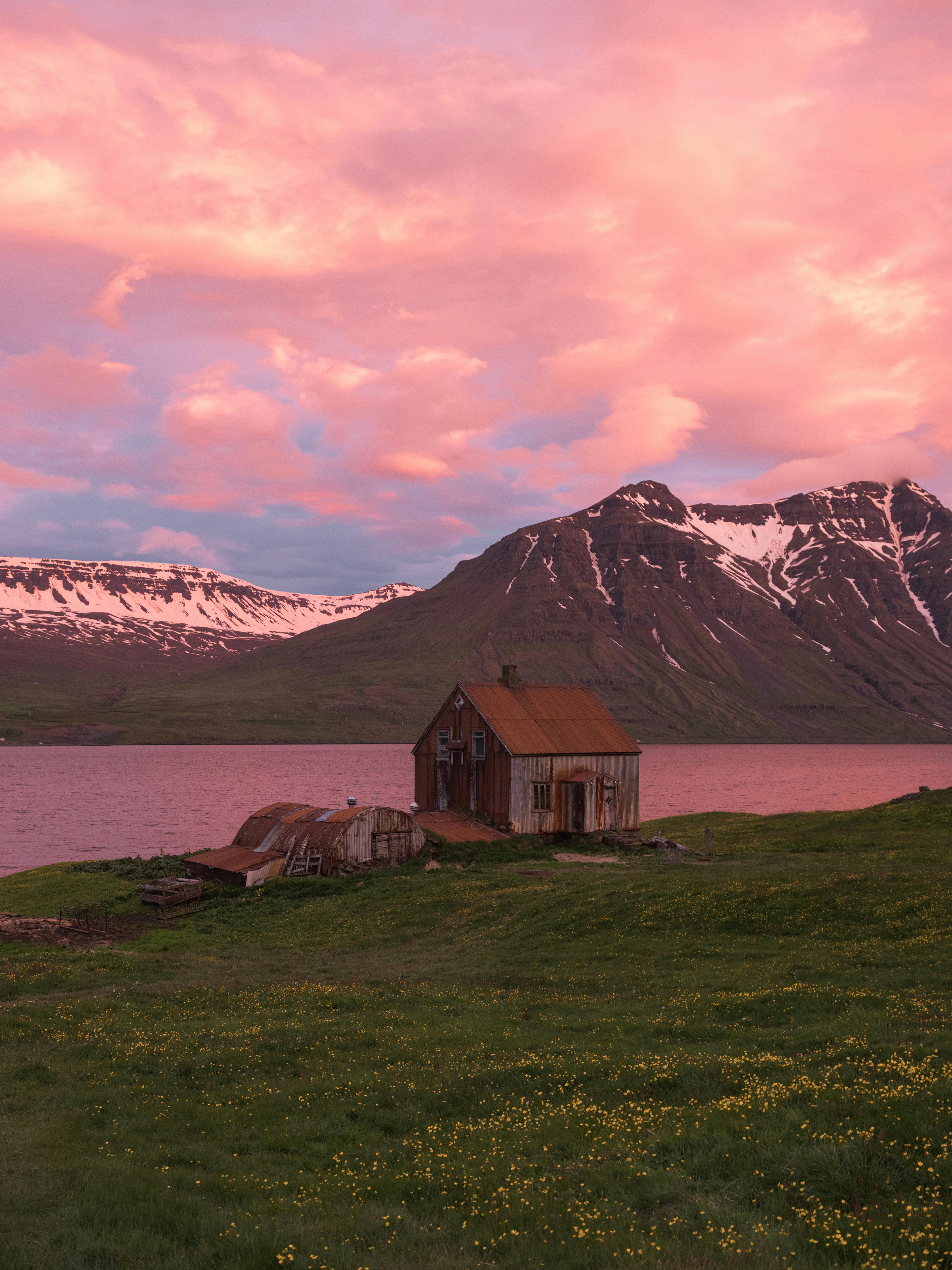 Spectacular idyllic landscape of snowy mountains and calm river located near grassy highland with old wooden house under beautiful purple sky