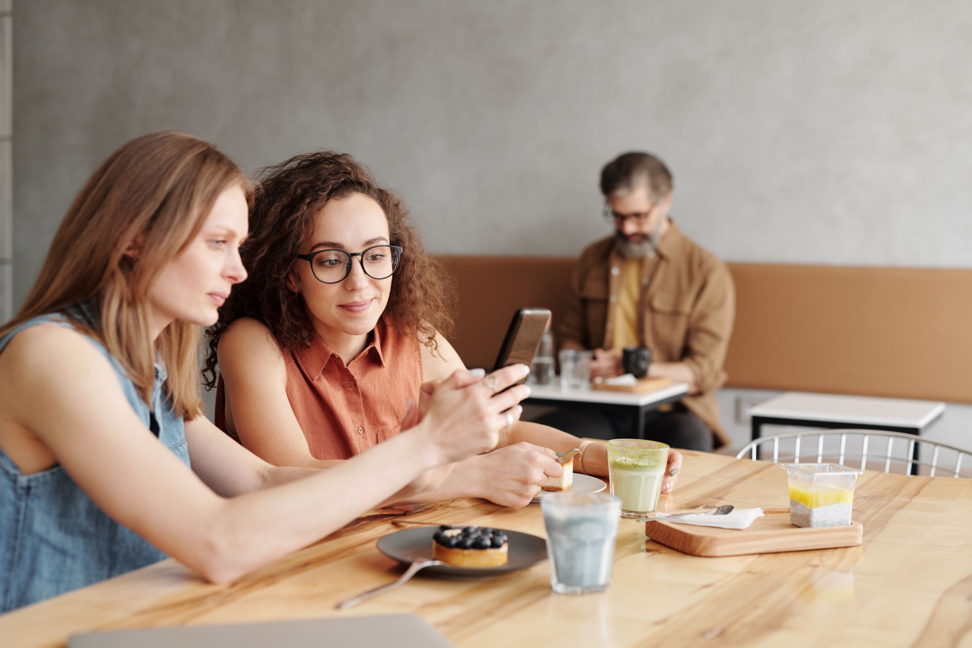 Two women in a cozy cafe, sharing drinks and using a smartphone, while a man enjoys his coffee in the background.
