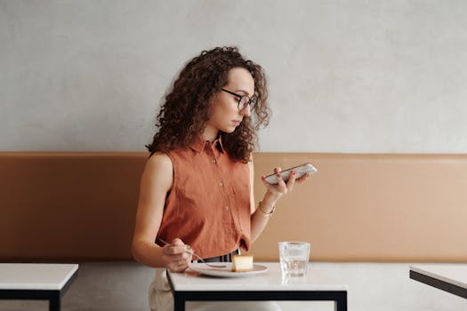 Woman with curly hair multitasks with smartphone and food in a cafe setting.