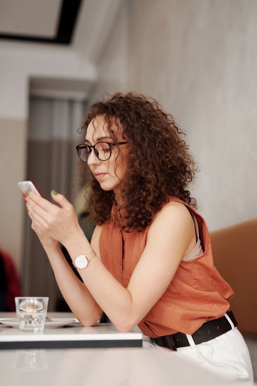 Free Woman with curly hair and eyeglasses using smartphone at cafe table, daytime. Stock Photo
