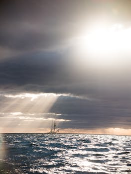 Lonely distant ship sailing in middle of wavy ocean against sunny rays falling through clouds