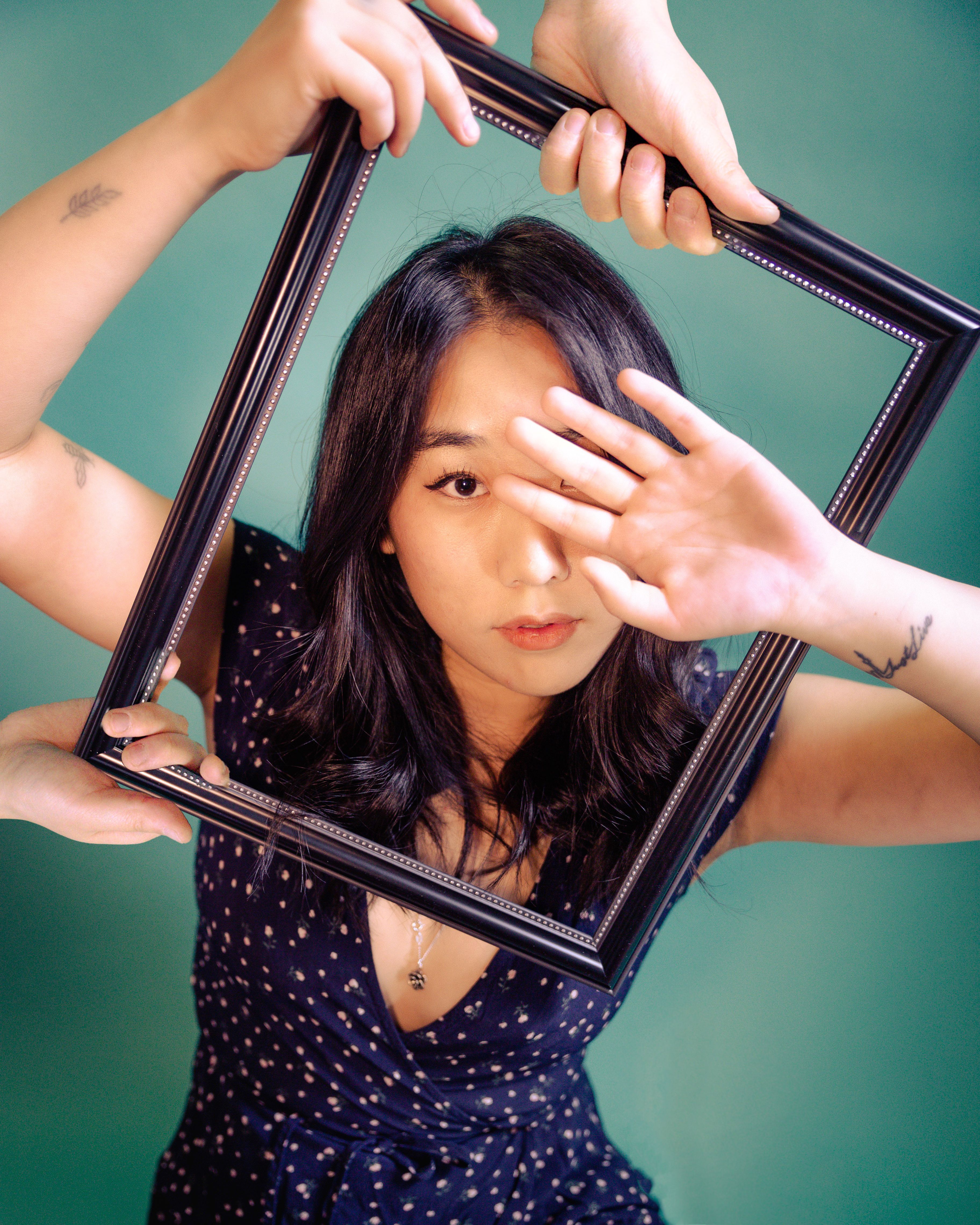 Artistic portrait featuring an Asian woman framed by hands against a teal background.