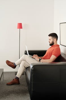 Bearded man in a polo shirt using a laptop on a black leather couch in a modern living room.