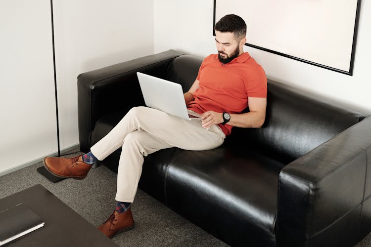 A Man In Red Polo Shirt And White Pants Sitting On Leather Couch While Using His Laptop