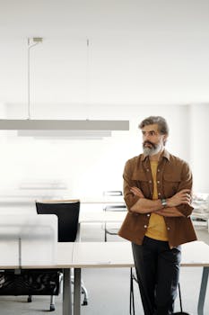 Bearded man in a modern office setting, arms crossed, showcasing contemporary professional style.
