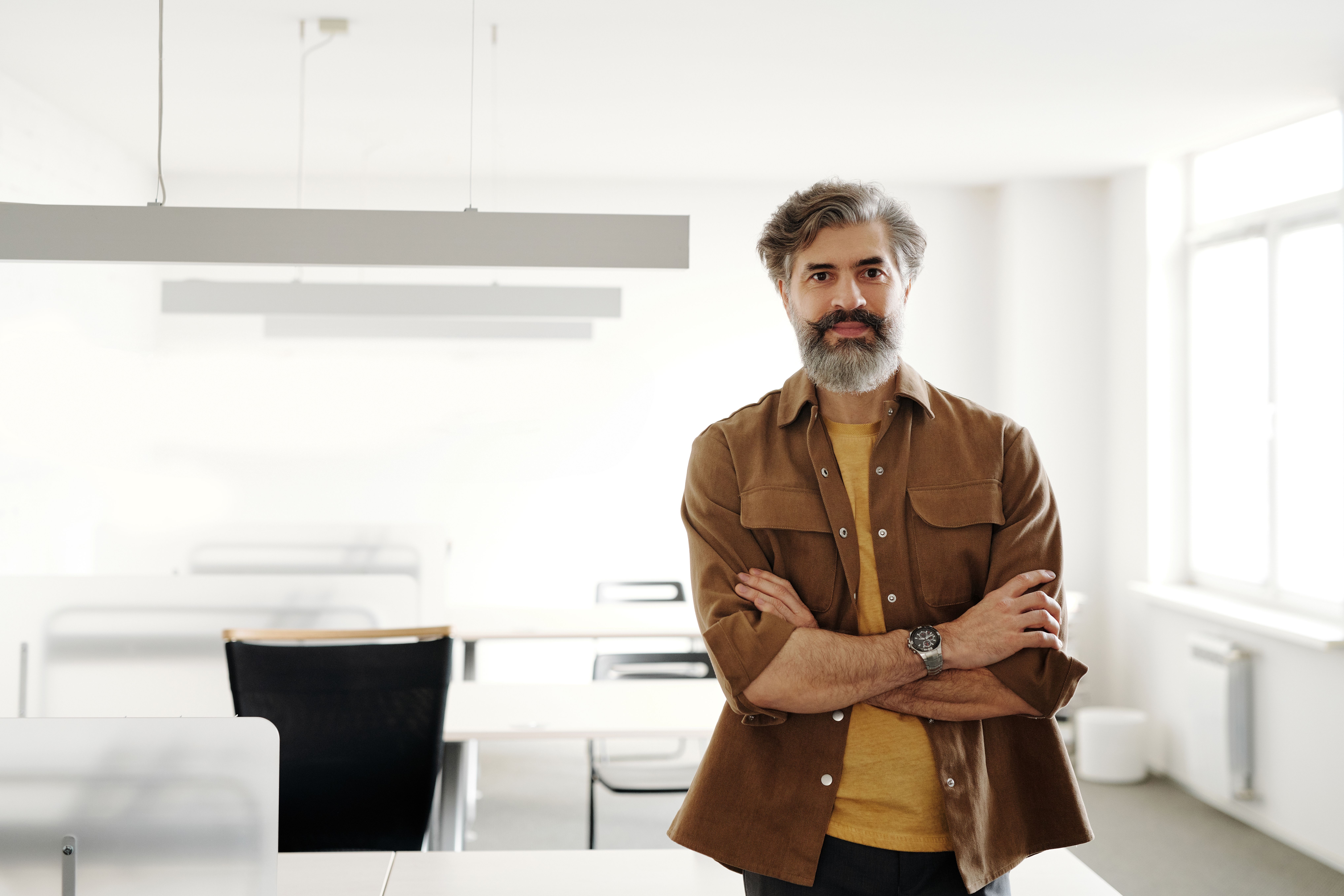A mature man with arms crossed standing confidently in a bright, modern office.