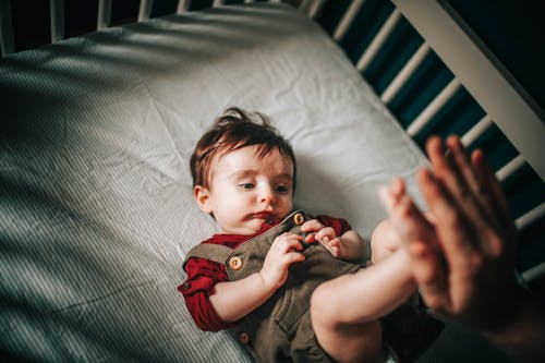 Free From above of crop anonymous parent touching feet of adorable little boy lying in crib on crumpled sheet while looking away at home Stock Photo