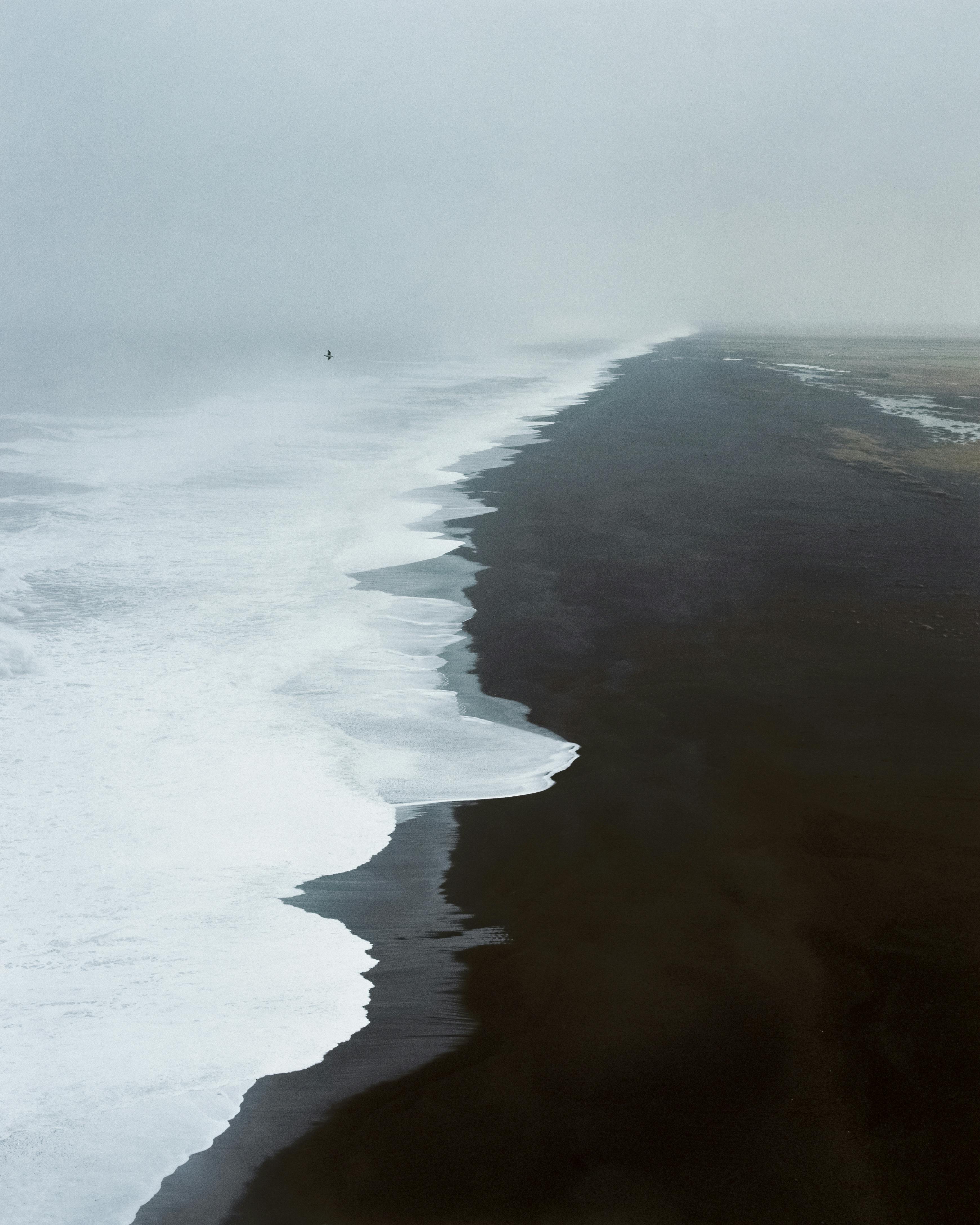 Drone view of foamy ocean waves rolling on black volcanic beach on overcast day