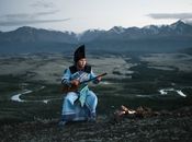 Full body of concentrated Mongolian male musician in national costume sitting near bonfire and playing traditional musical instrument against picturesque mountains