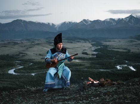 Full body of concentrated Mongolian male musician in national costume sitting near bonfire and playing traditional musical instrument against picturesque mountains