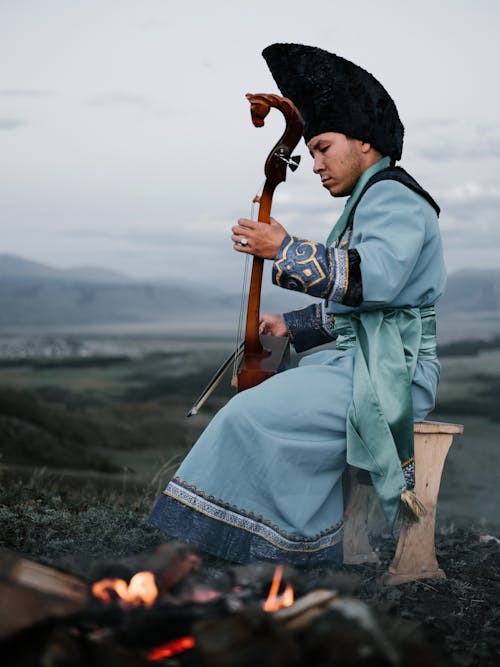 Side view of thoughtful Mongolian male musician in traditional costume sitting on stool in valley and playing morin khuur musical instrument