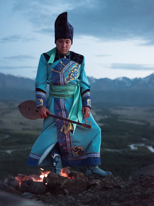 Thoughtful young Mongolian male in traditional clothes looking at bonfire while standing in valley with folk musical instrument
