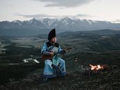 Tibetan young man playing lute sitting against mountains in twilight