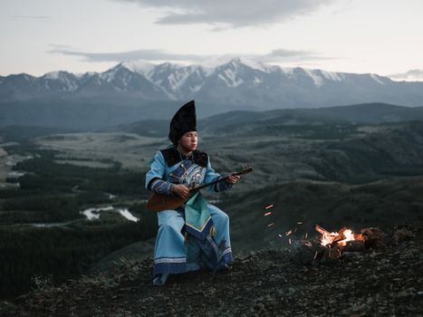 Tibetan young man playing lute sitting against mountains in twilight