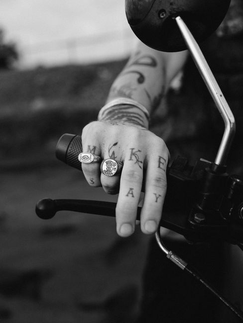 A close-up black and white shot of a tattooed hand gripping a motorcycle handlebar, adorned with rings.