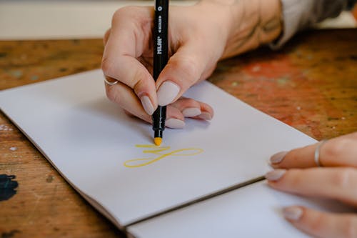Crop unrecognizable female designer practicing calligraphy while writing letters in blank album using yellow felt pen while sitting at wooden table