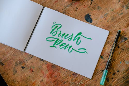 Top view of rectangular paper album with BRUSH PEN title written with green felt pen and original letter font on wooden desk with ink spots in workshop