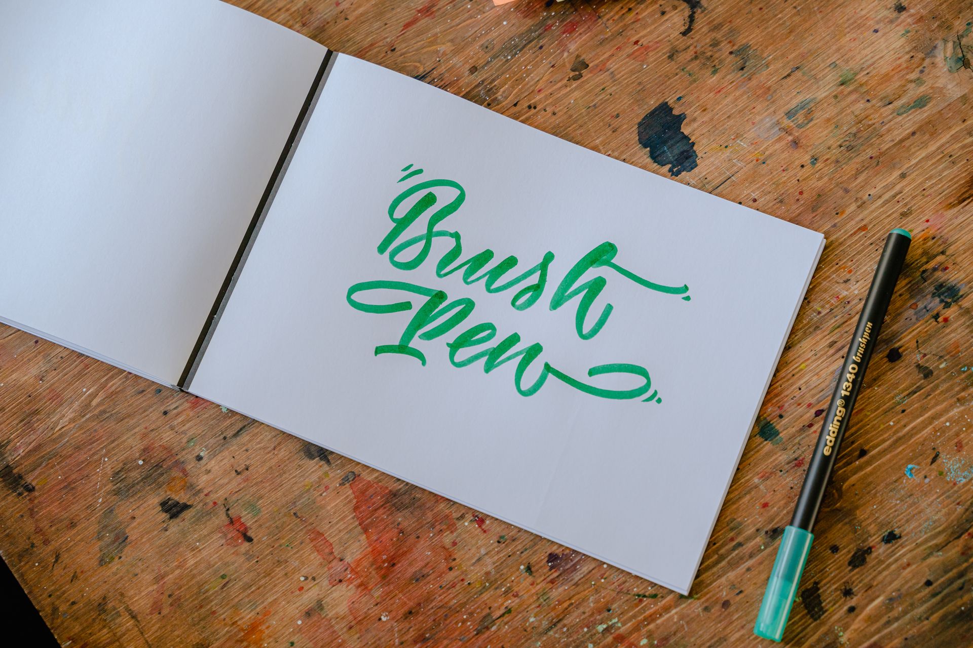 Top view of rectangular paper album with BRUSH PEN title written with green felt pen and original letter font on wooden desk with ink spots in workshop