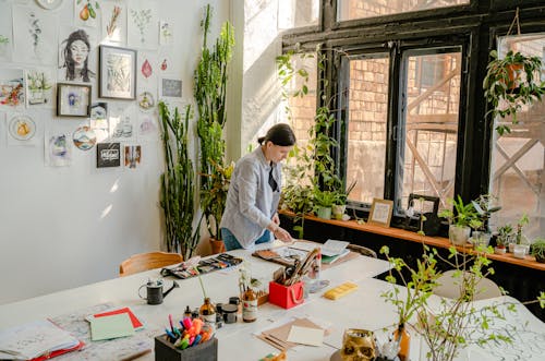 Side view of female designer creating drawings at desk with collection of felt pens and papers near wall with artworks and plants on windowsill in daylight