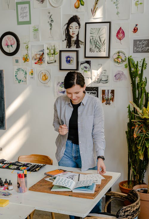 Cheerful young female designer in jeans standing near wall with pictures and table with stationery while choosing drafts with letter design in daylight