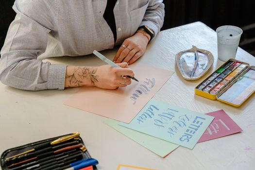 From above of crop tattooed female designer using marker white writing word with creative letter design on multicolored papers while sitting at table with artist watercolor palette