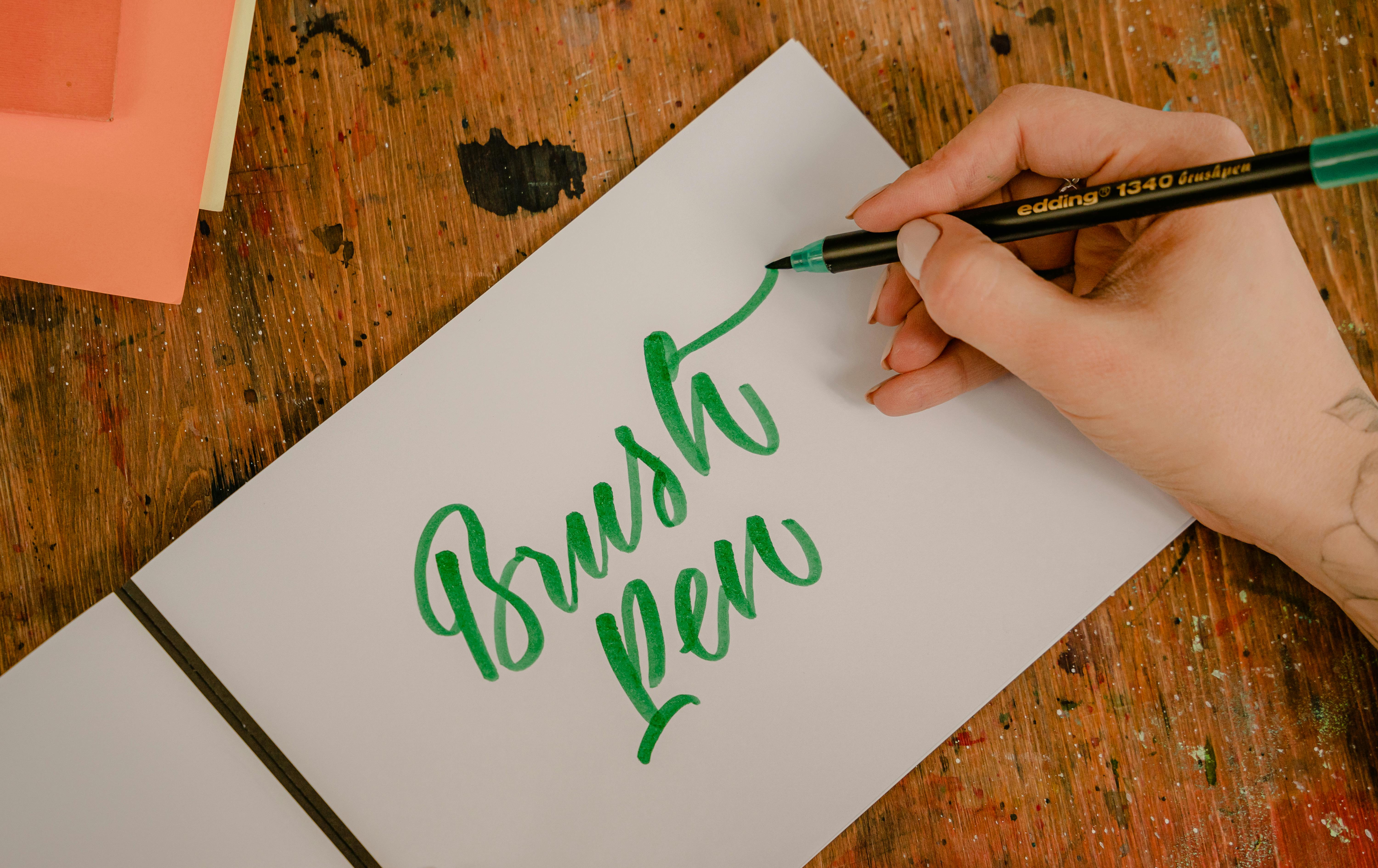 Top view of crop anonymous female designer writing BRUSH PEN title using felt pen and creative letter font while sitting at wooden table with ink spots on surface