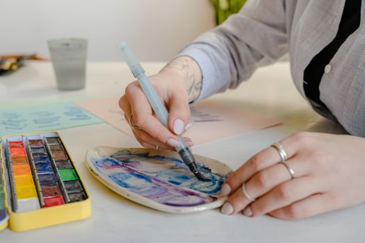 Crop faceless tattooed female designer using paint brush and palette while mixing blue color tones and sitting at table with sheets of paper illustrating different letter fonts