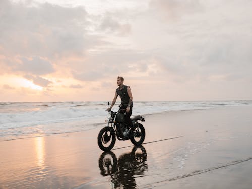 Tattooed biker riding modern motorcycle reflecting on sand of ocean beach near foamy waves under picturesque cloudy sky at sundown