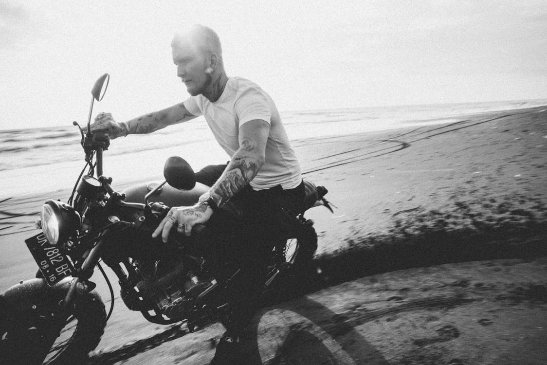 Brutal bearded hipster in t shirt  and wristwatch riding motorbike with headlight on passing on sandy sea coast with trace in back lit