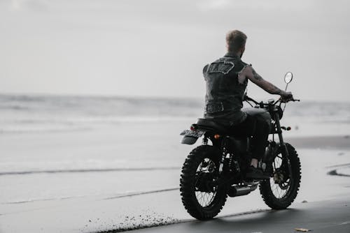 Back view of unrecognizable tattooed rider in sleeveless shirt riding modern bike on sandy sea shore leaving trace under sky with clouds