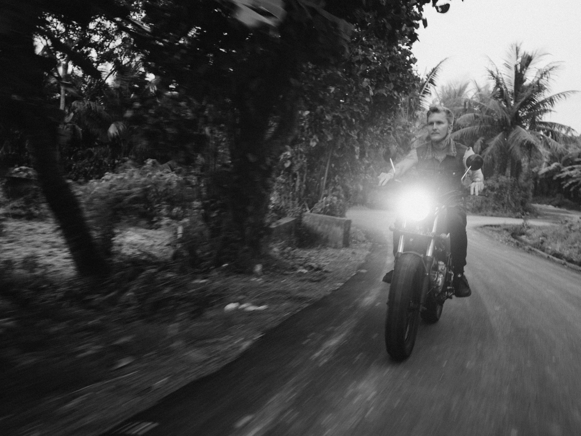 Black and white photo of a man riding a motorcycle on a winding road in a lush outdoor setting.