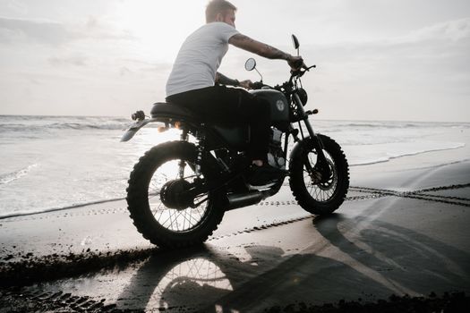 From below side view of unrecognizable tattooed guy in t shirt riding motorbike on sand beach leaving traces and creating splash effect near sea in evening in back lit