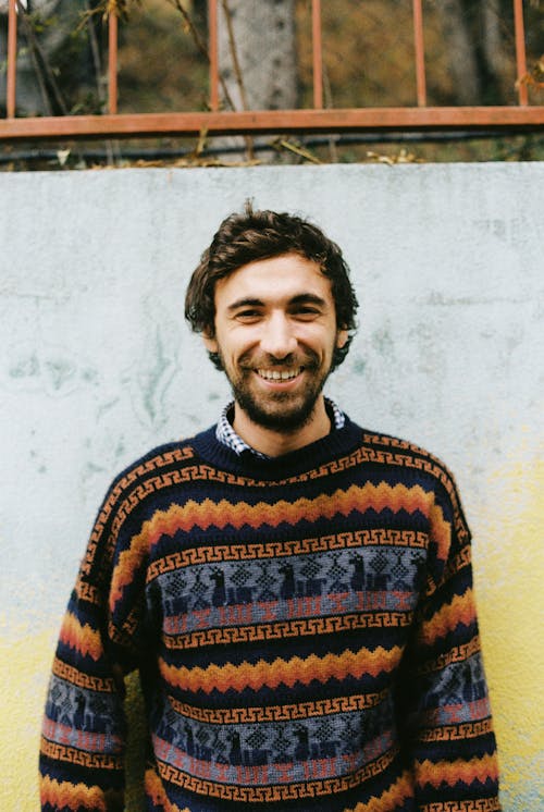 Happy adult bearded male with toothy smile in knitted sweater standing near colorful shabby wall with metal fence above in daylight and looking at camera