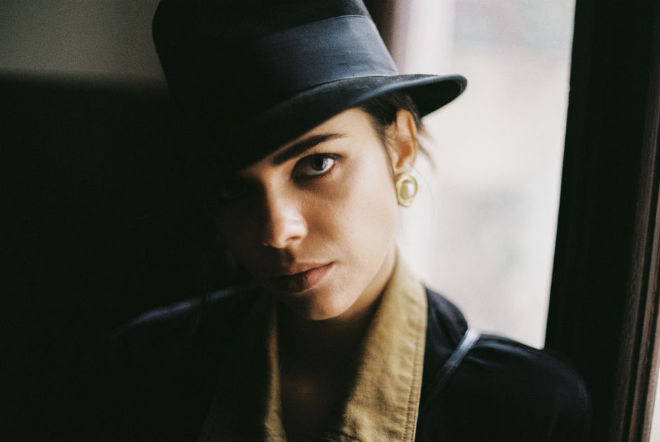 Young lady with brown eyes in golden earrings standing in black hat while showing half of face near window in daylight and looking at camera