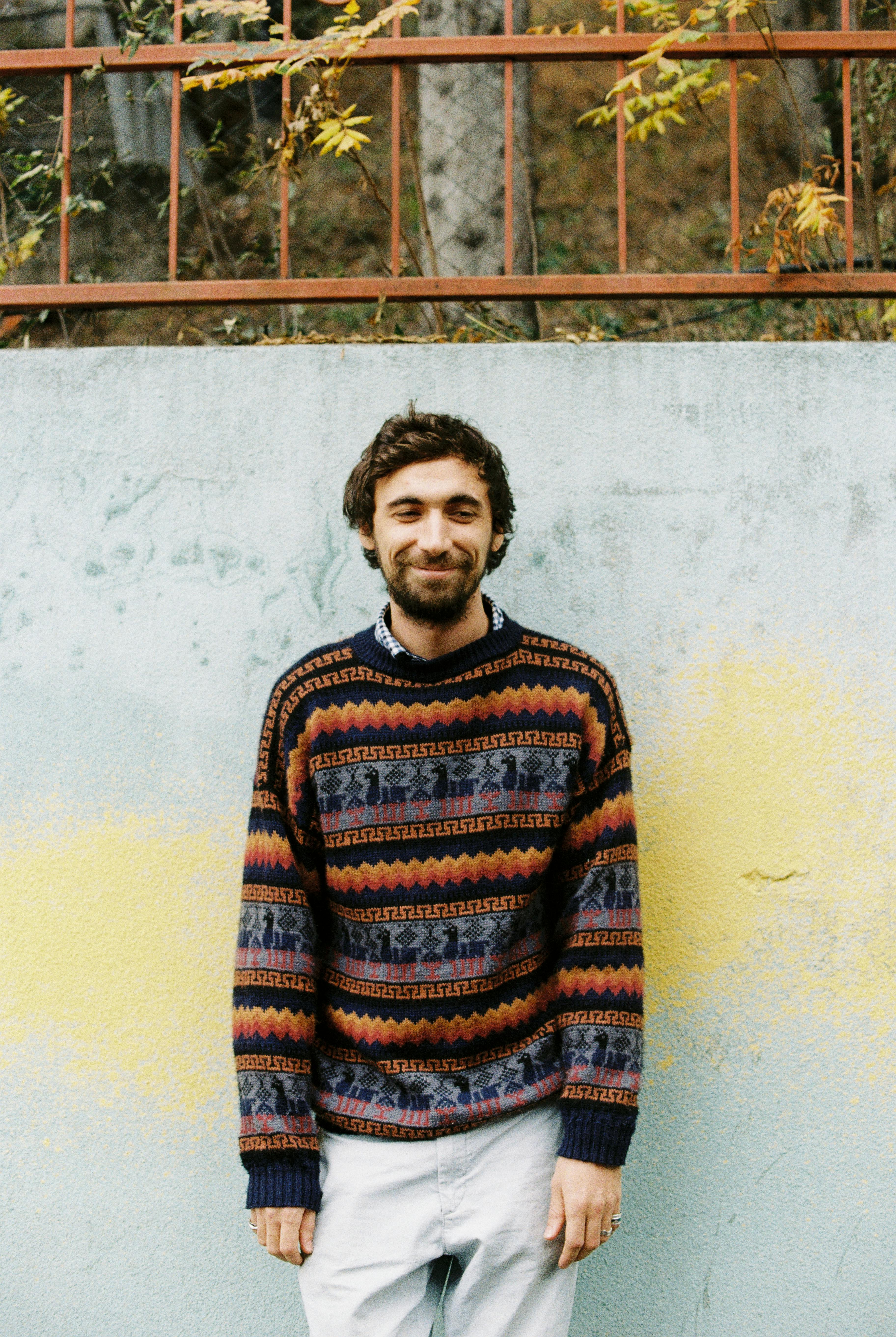 Cheerful adult slender bearded male in knitted sweater and jeans looking at camera while standing near shabby wall with metal fence above and tree trunk with faded leaves in fall