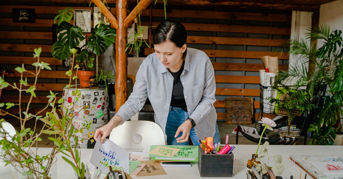 Woman arranging calligraphy materials in a plant-filled studio setting, emphasizing creativity.