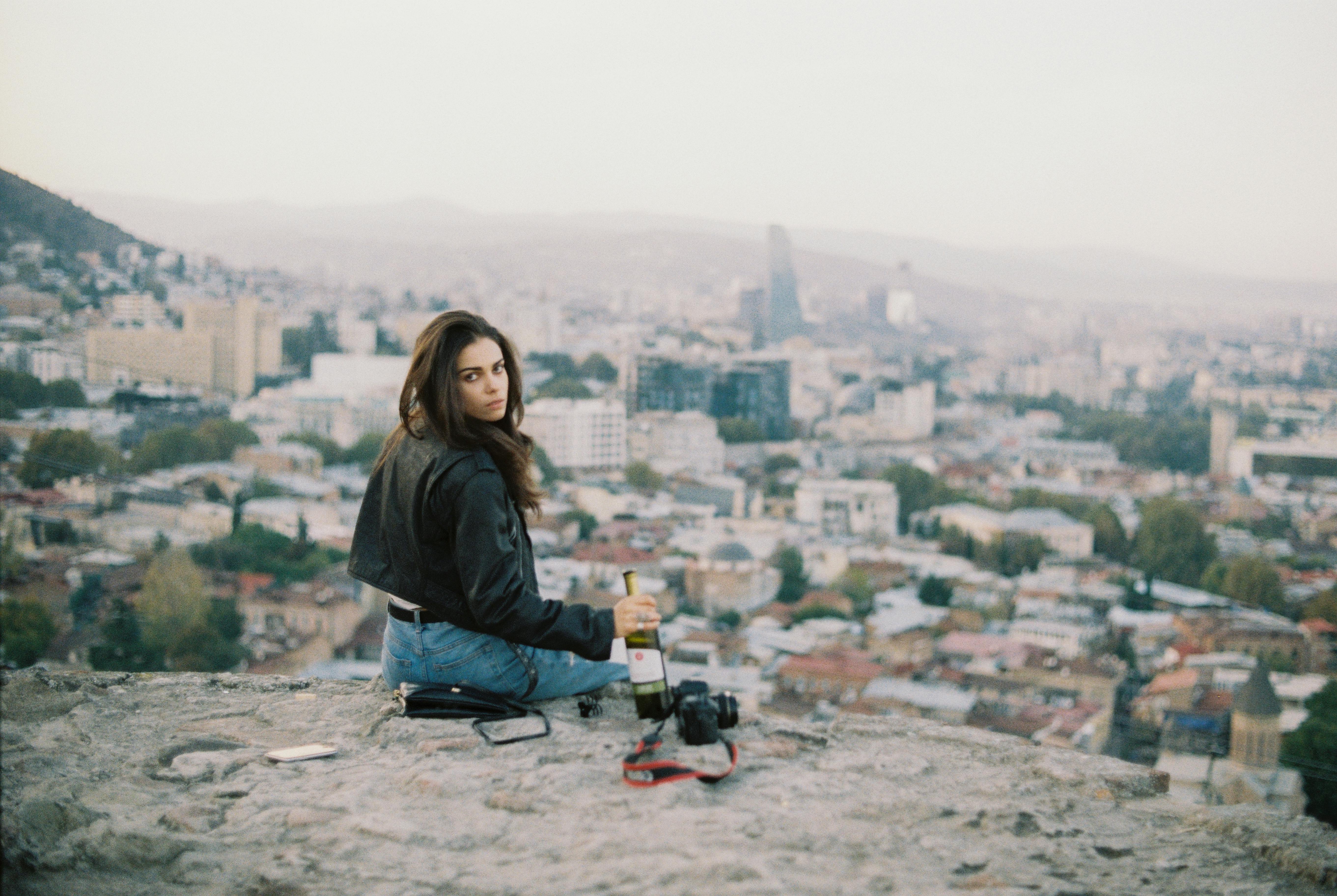 Serious young female in casual clothes sitting above city on parapet and holding bottle of wine in hand while looking at camera