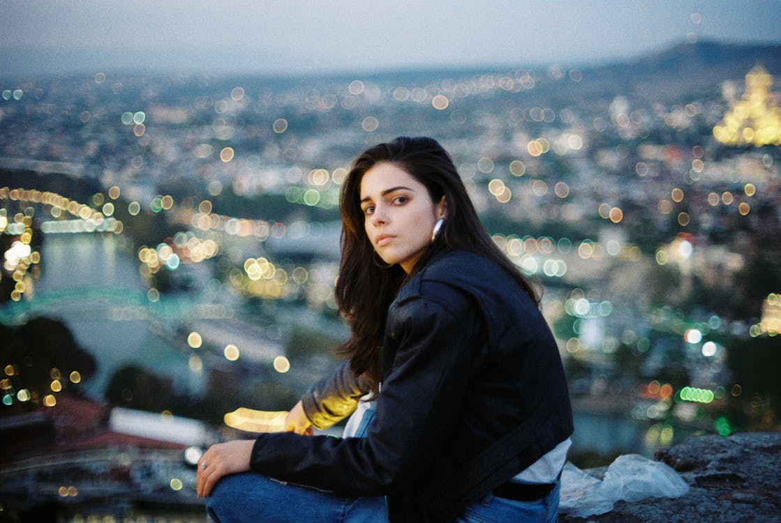 Free Emotionless young female in black jacket and jeans sitting on rooftop above city street in evening time and looking at camera Stock Photo