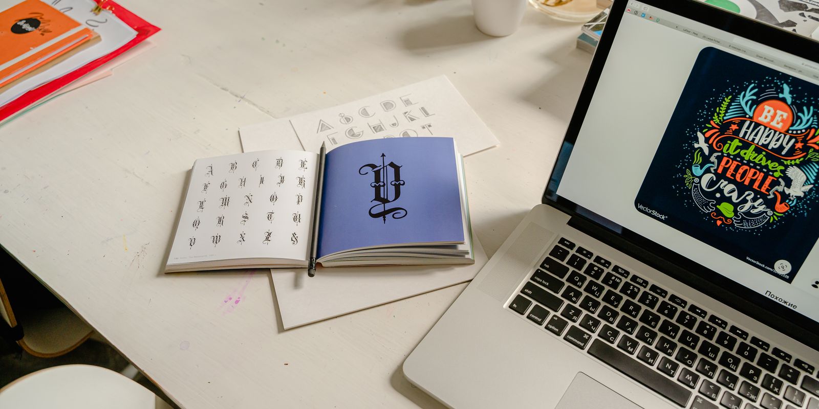 An open calligraphy type specimen book next to a laptop showing a colorful lettering design