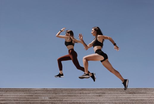 Two women in active wear leap energetically on stairs against a clear blue sky.