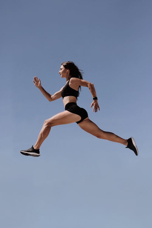 Woman in Black Sports Bra and Black Shorts Jumping on Air Under Blue Sky