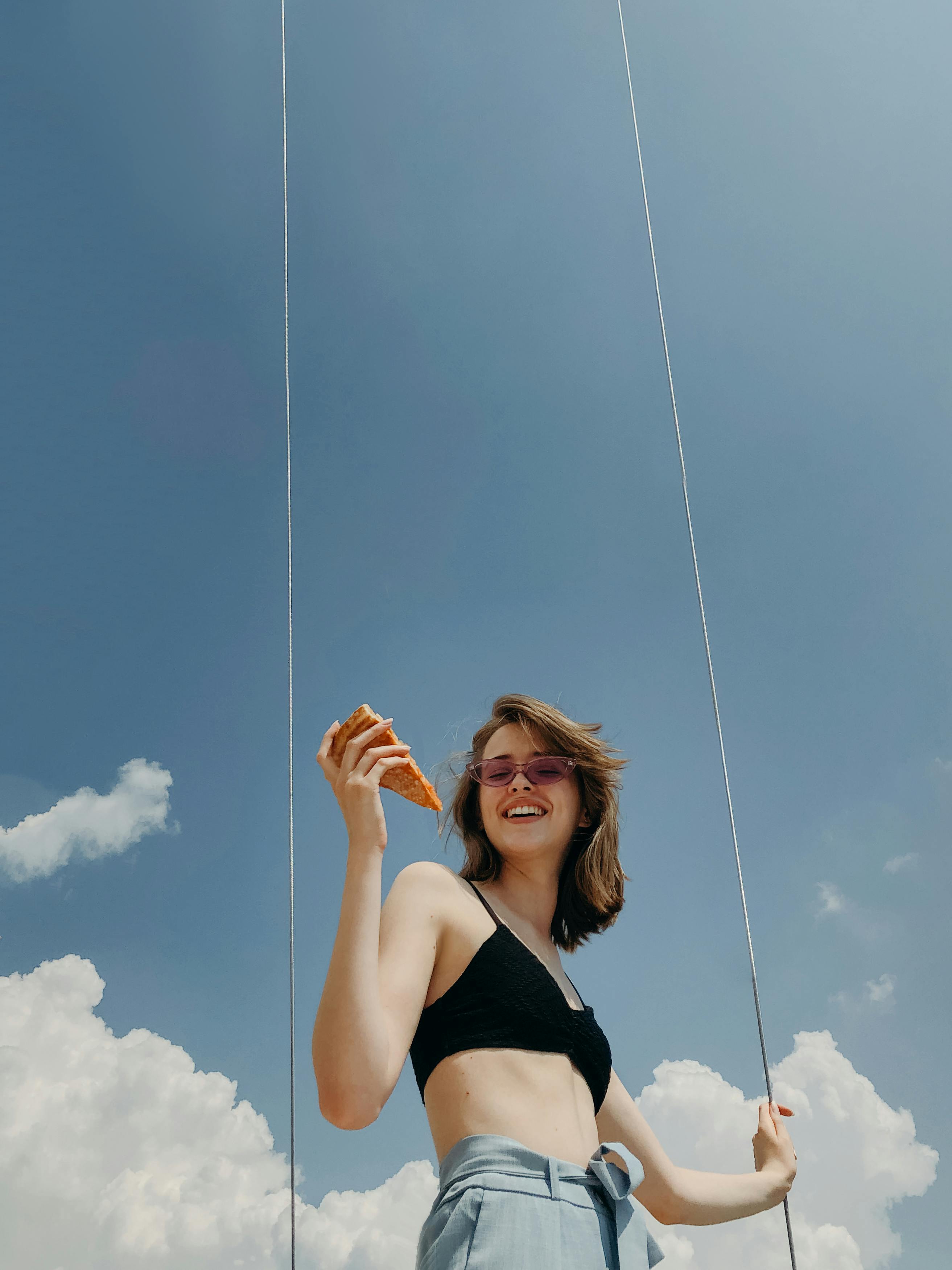 Smiling woman in crop top with sunglasses holding a pizza slice against a clear blue sky.