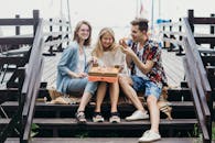 三位年轻的朋友在户外的木台阶上分享一个比萨饼，微笑着捕捉有趣的瞬间